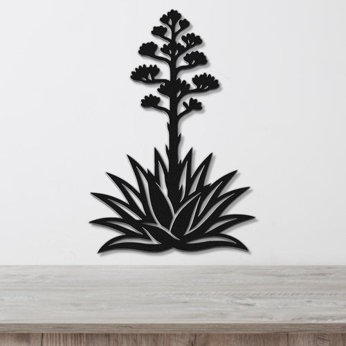 Black metal wall art in the silhouette of a blooming agave plant with cutout leaves and flower stalk, mounted on a white wall above a wooden surface.