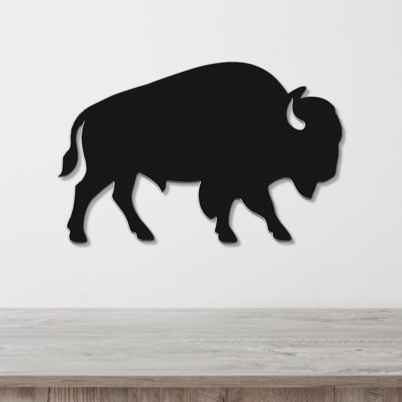 Black metal buffalo wall art silhouette with horn and tail cutouts, mounted on a white wall above a light wood surface.