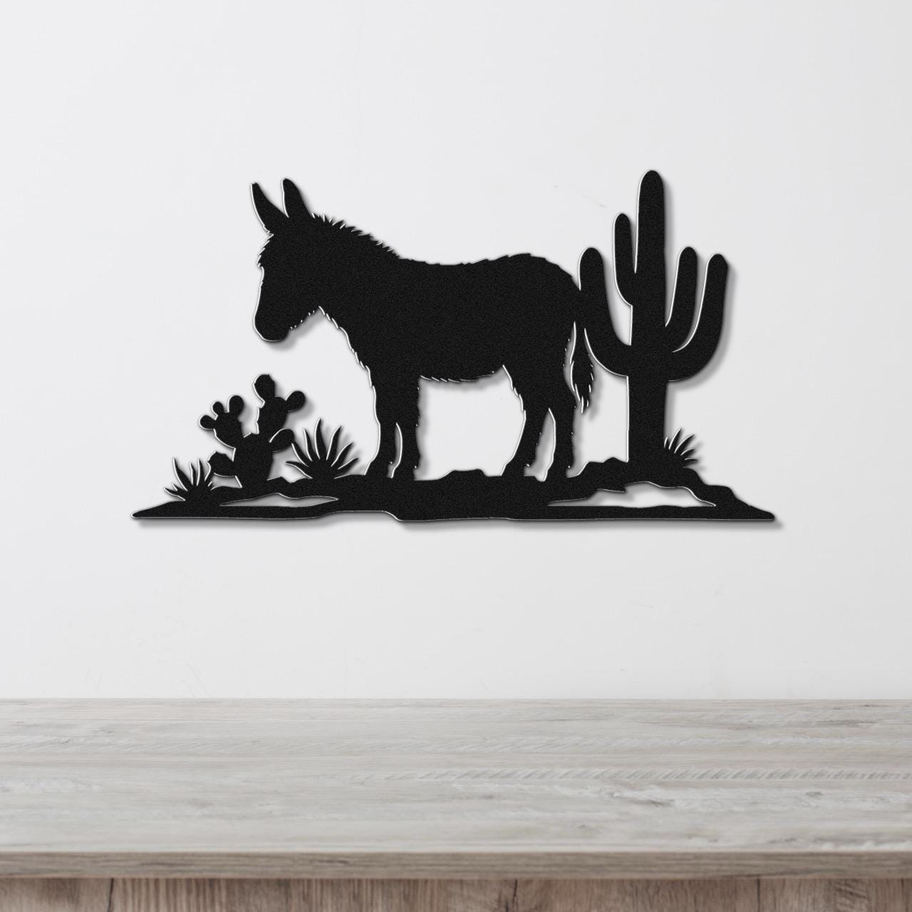 Black metal wall art silhouette of a burro and cacti, mounted on a white wall above a wooden surface.