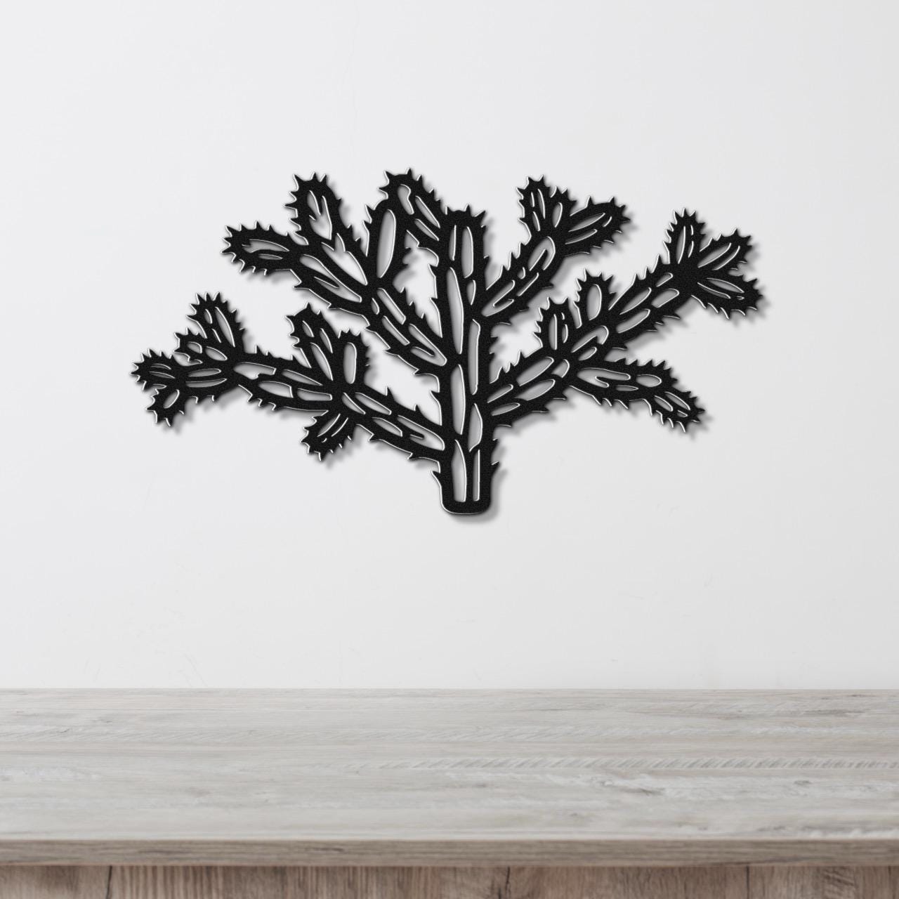 Black metal wall art cutout of a cholla cactus with intricate branching and spiny segments, mounted on a white wall above a light wood surface.