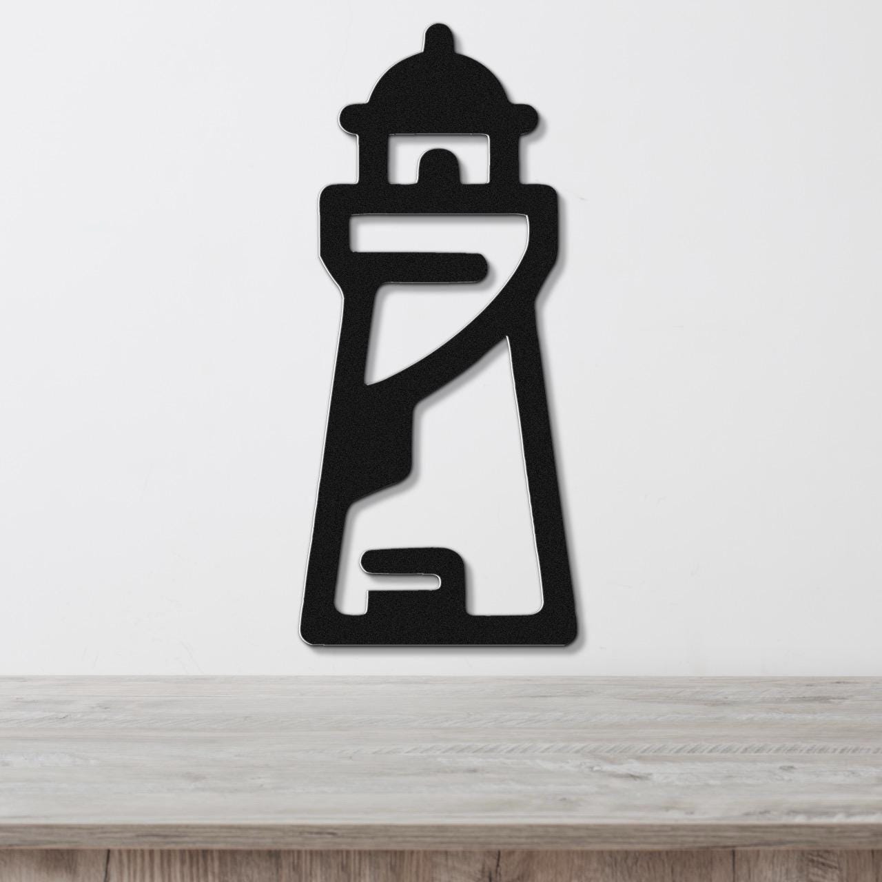Black metal lighthouse wall art with cutout details, mounted on a white wall above a wooden surface.