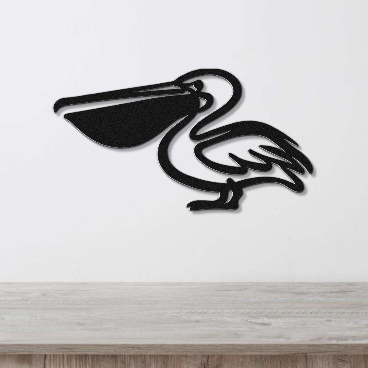 Black metal pelican wall art with a cutout silhouette design, mounted on a white wall above a light wood surface.