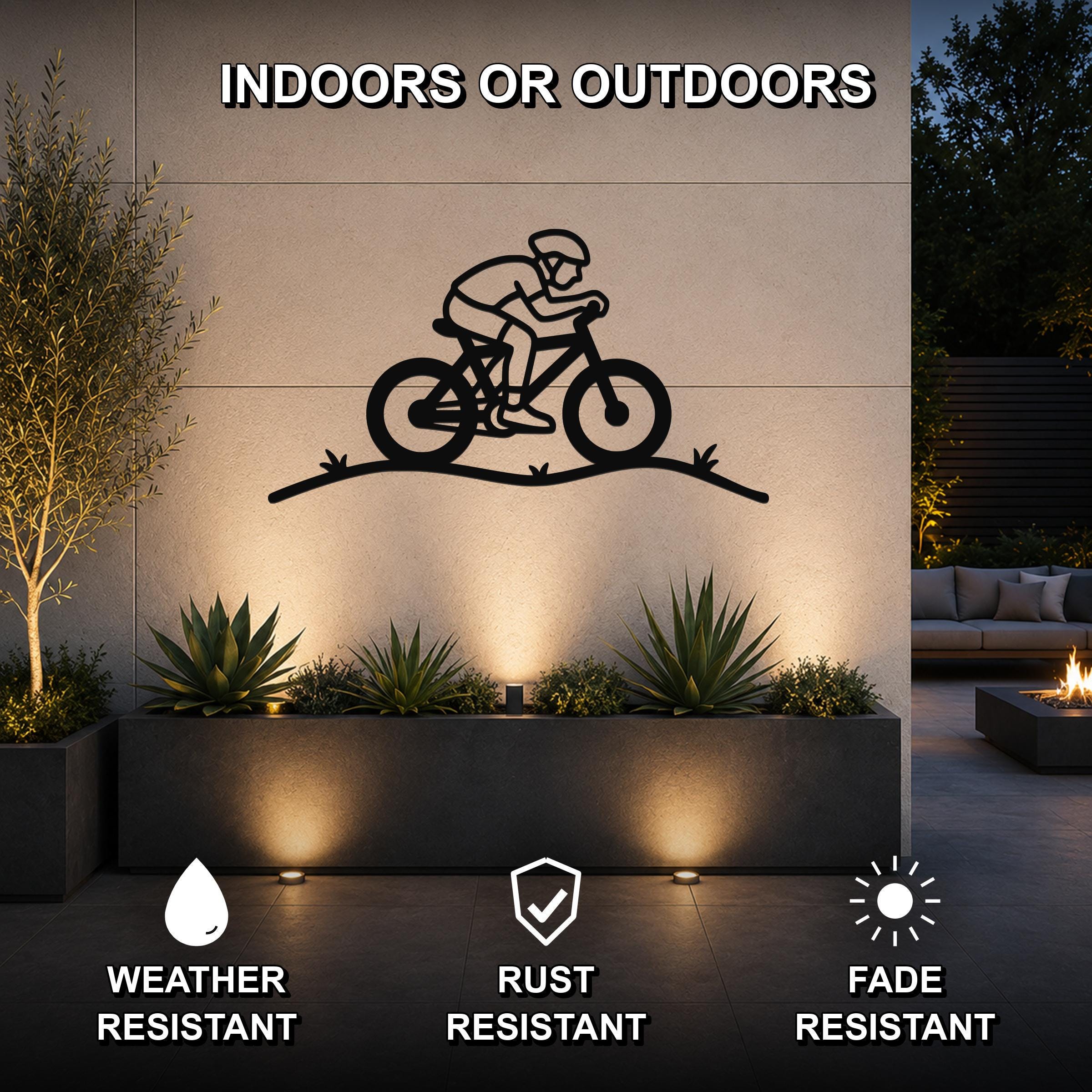 Black metal wall art featuring a cyclist silhouette on a hill, mounted on an outdoor wall above a planter in a patio setting.