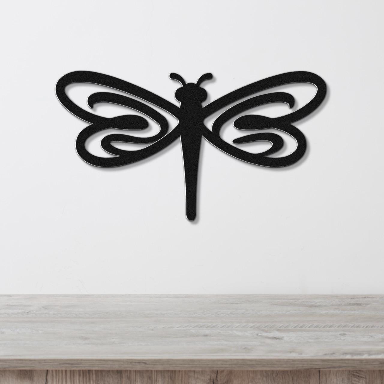 Black metal dragonfly wall art with flowing wing cutouts, mounted on a white wall above a light wood surface.