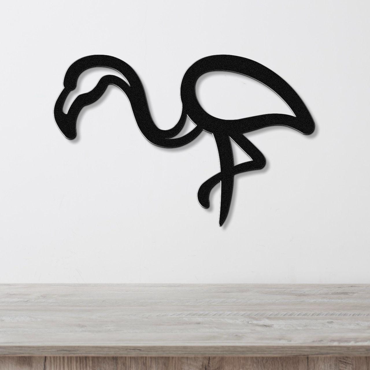 Black metal flamingo wall art with a bold silhouette design, mounted on a white wall above a light wood surface.