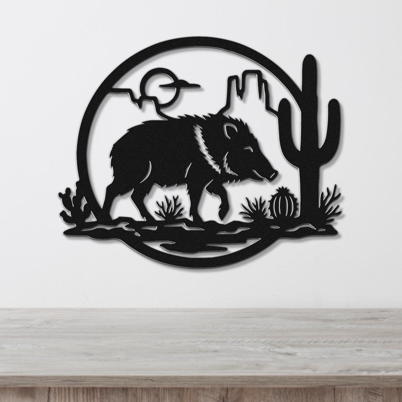 Black metal wall art silhouette of a javelina in a desert scene with cactus and mountains, mounted on a white wall above a wooden surface.