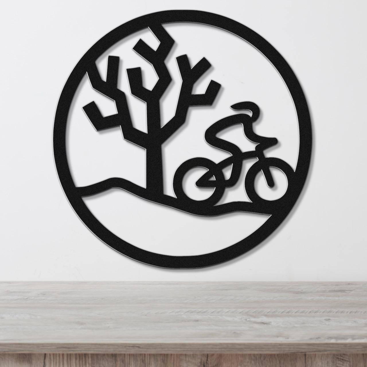 Black metal circular wall art with a cyclist in motion and a tree silhouette, mounted on a white wall above a wooden surface.