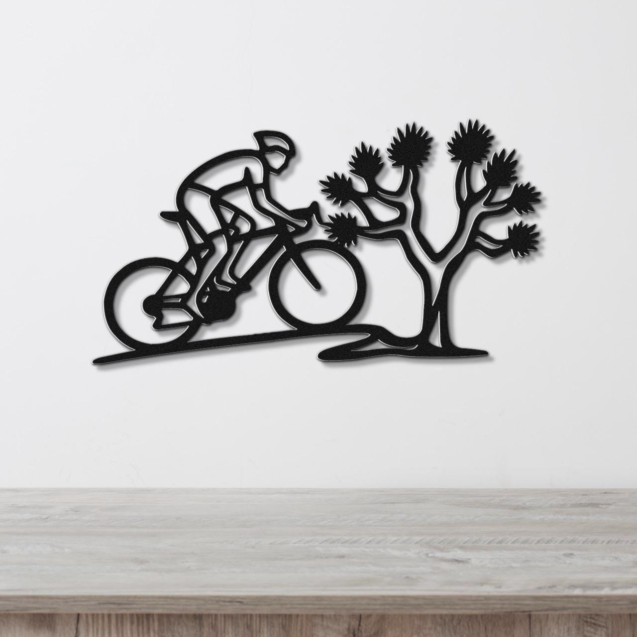 Black metal wall art cutout of a cyclist riding past a Joshua tree, mounted on a white wall above a wooden surface.