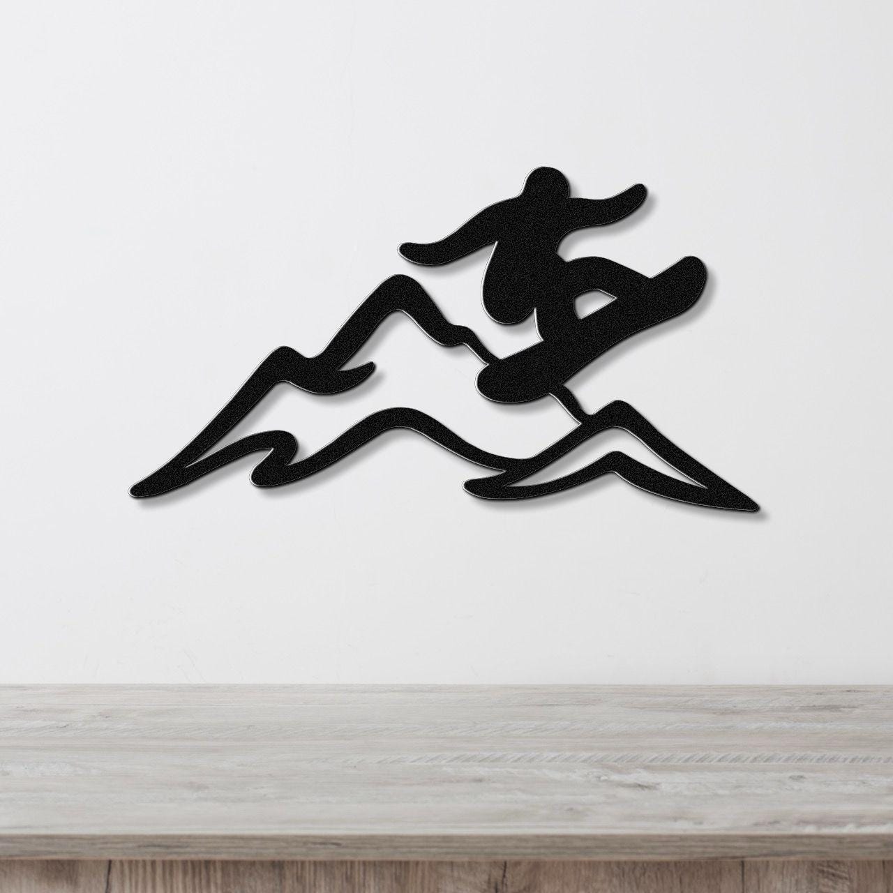 Black metal wall art silhouette of a snowboarder in motion over mountain peaks, mounted on a white wall above a wooden surface.