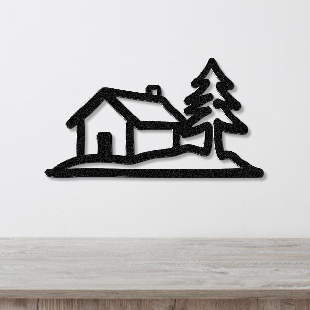 Black metal wall art cutout of a cabin and pine tree silhouette, mounted on a white wall above a light wood counter.