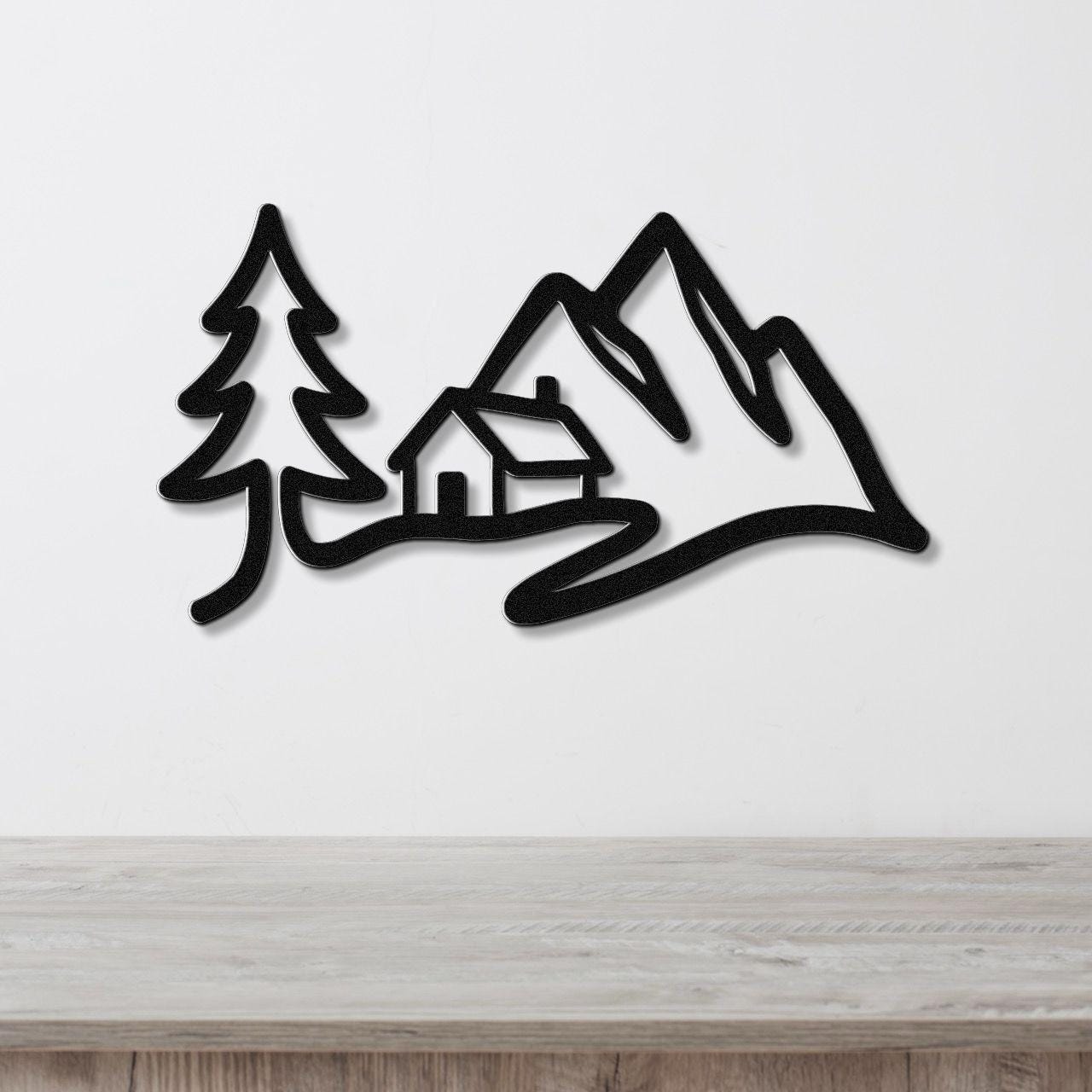Black metal wall art silhouette of a mountain cabin scene with a pine tree and peaks, mounted on a white wall above a wooden surface.