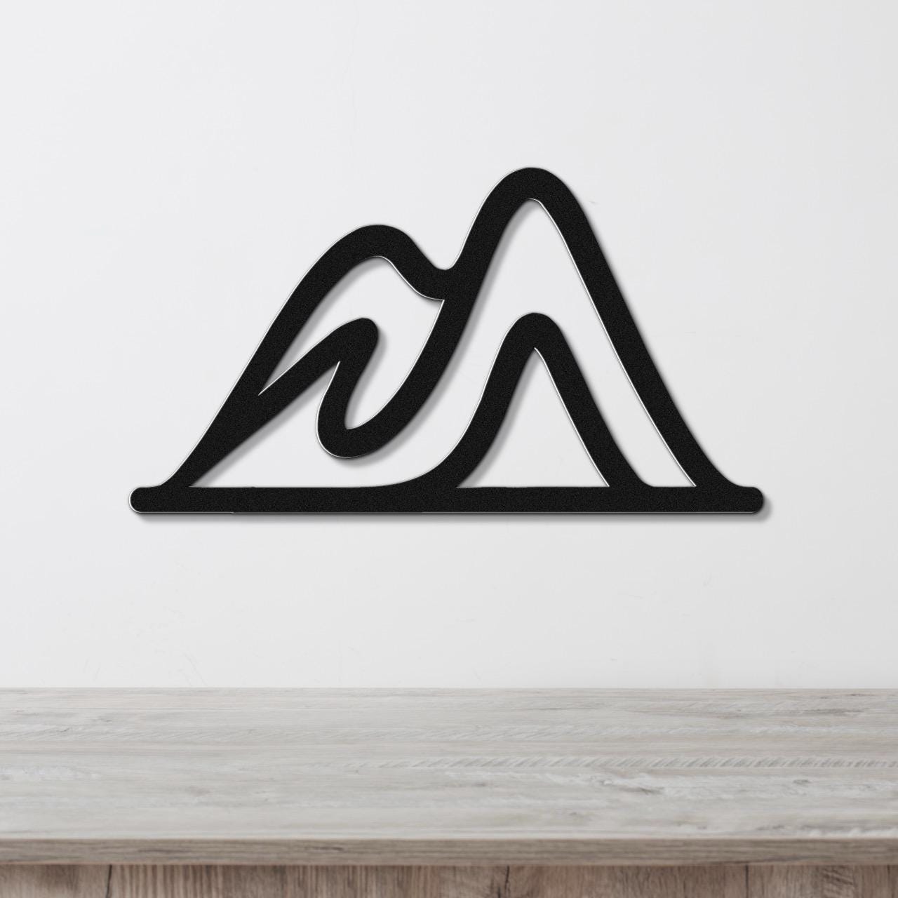 Black metal wall art with a mountain silhouette design, featuring layered peaks and internal cutouts, mounted on a white wall above a wooden surface.