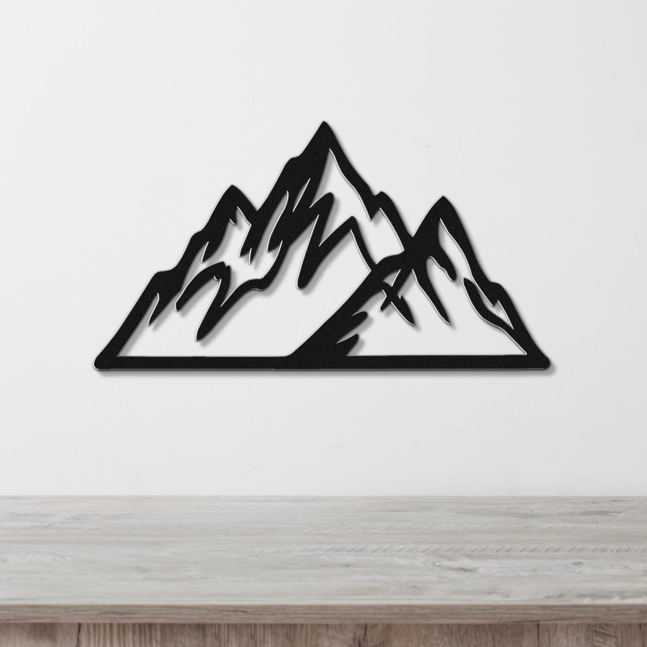 Black metal mountain wall art with layered peak cutouts, mounted on a white wall above a light wood surface.