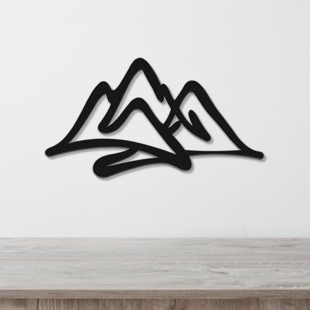 Black metal wall art with a layered mountain silhouette design, mounted on a white wall above a light wood surface.