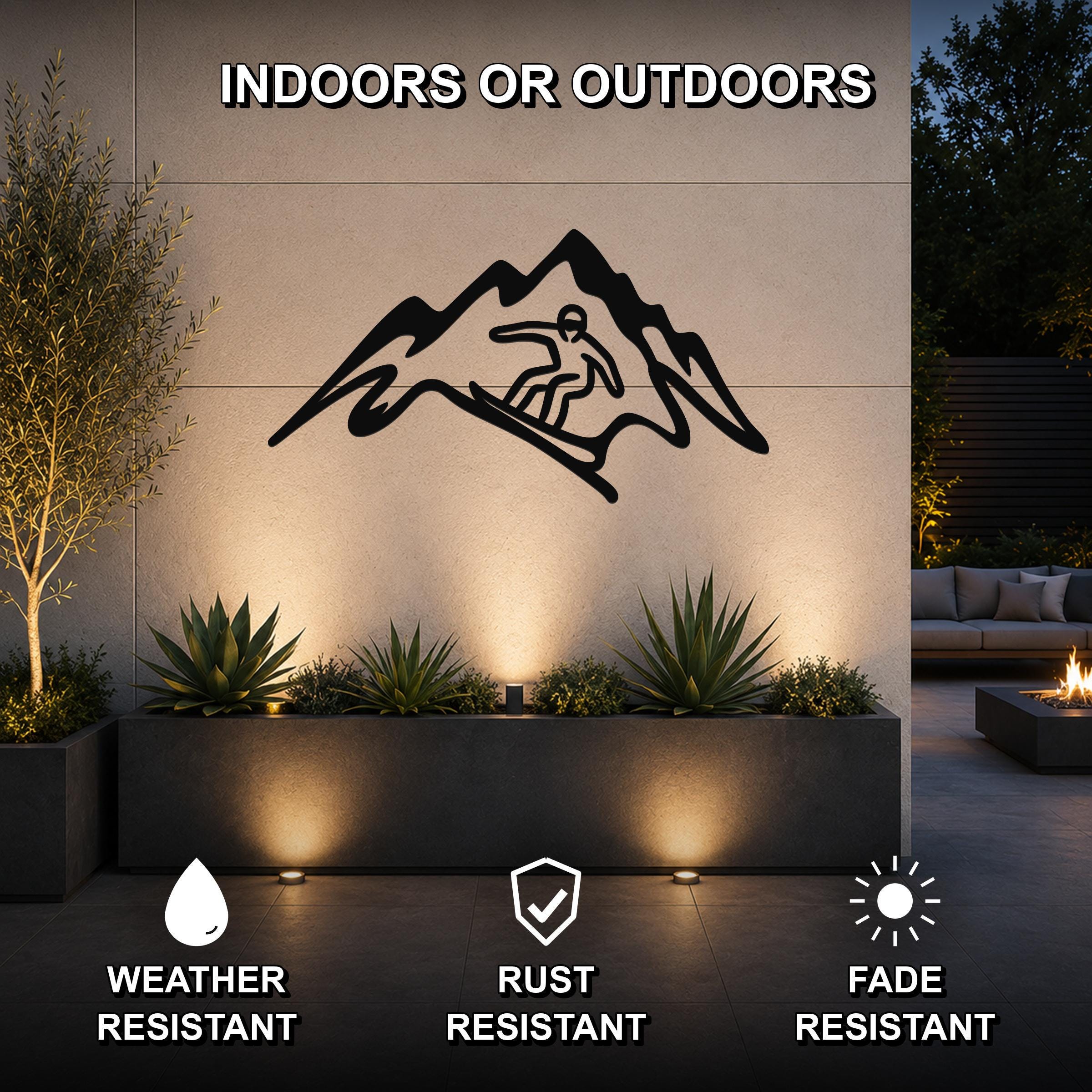 Black metal wall art featuring a snowboarder silhouette within a mountain cutout, mounted on an outdoor wall above a planter in a patio setting.