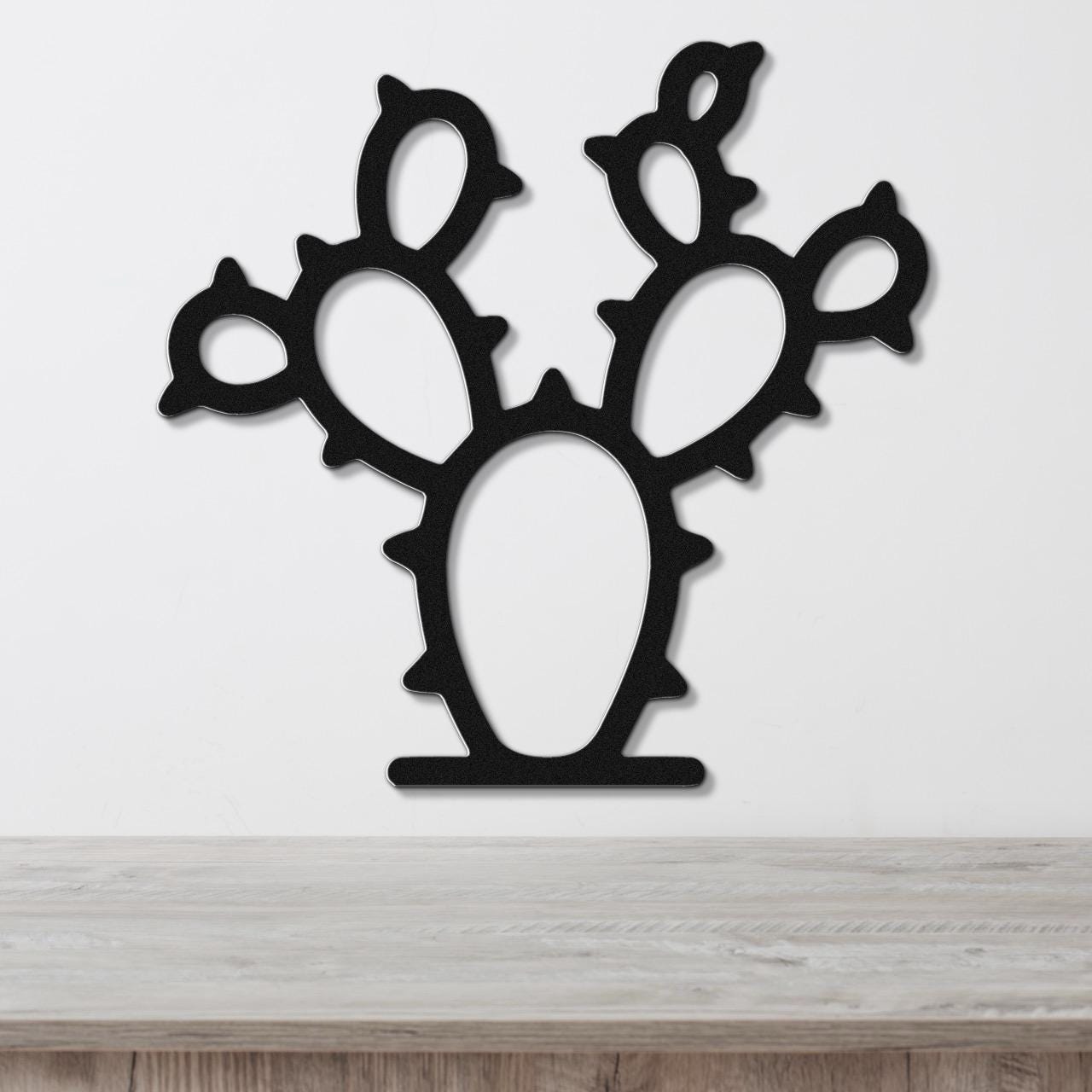 Black metal wall art cutout of a prickly pear cactus with multiple pads and spines, mounted on a white wall above a light wood surface.