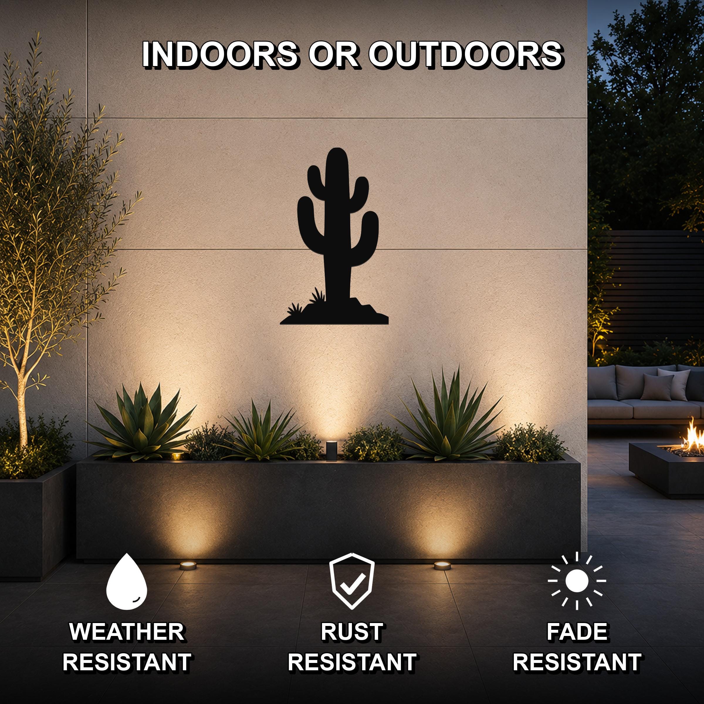 Black metal saguaro cactus wall art silhouette mounted on an outdoor wall above a planter, illuminated by uplights in a modern patio setting.