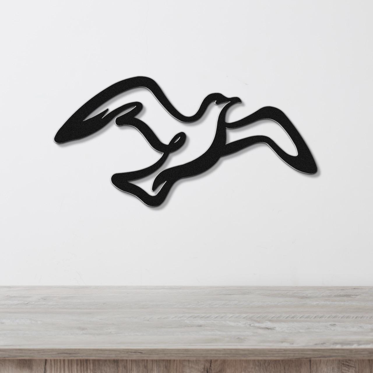 Black metal seagull wall art with flowing wing cutouts, mounted on a white wall above a light wood surface.