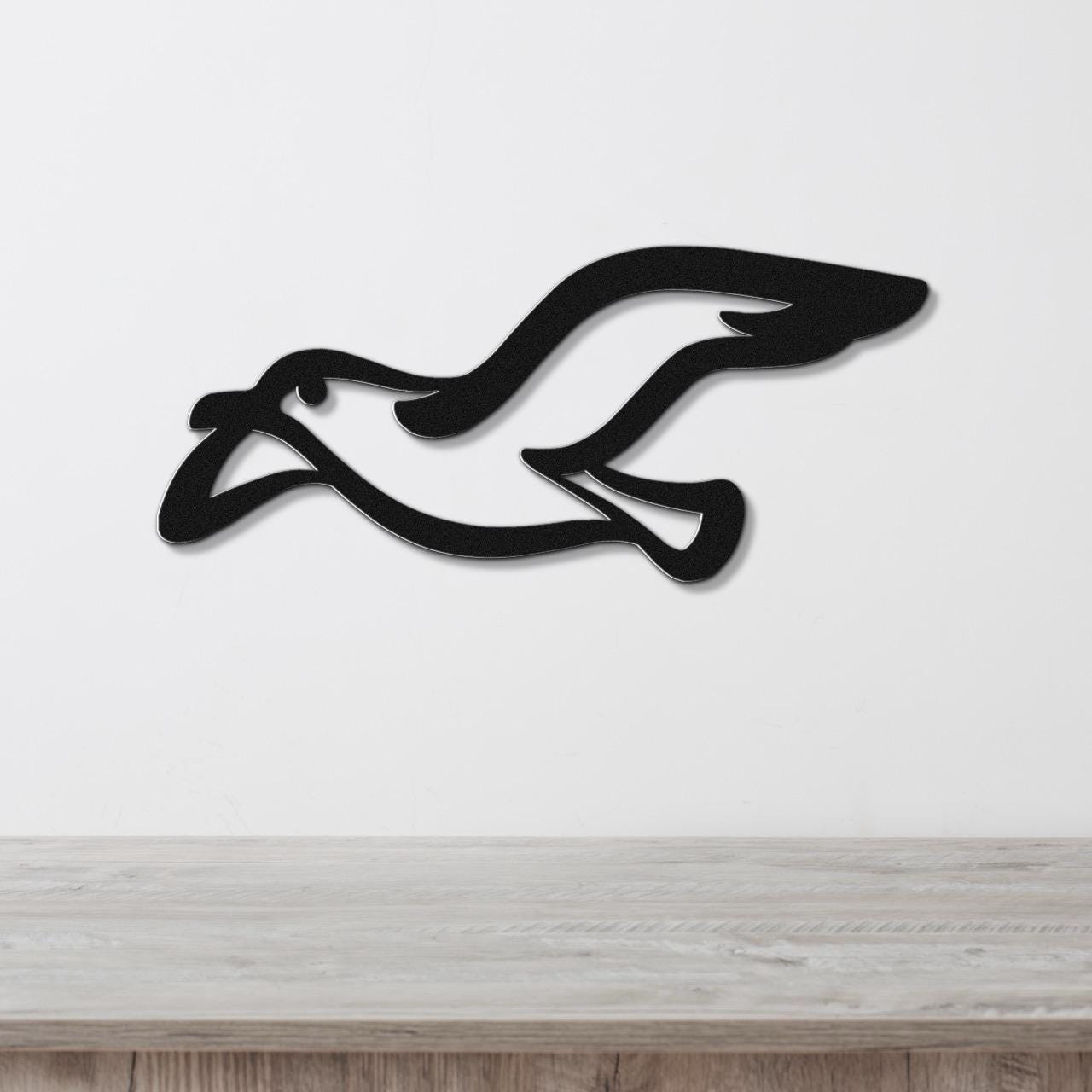 Black metal seagull wall art with a sleek silhouette design, mounted on a white wall above a light wood surface.