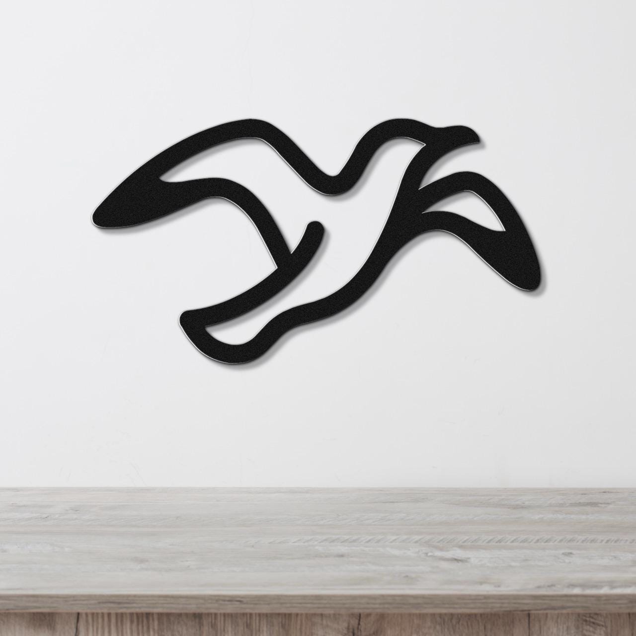 Black metal seagull wall art with flowing motion lines, mounted on a white wall above a light wood surface.