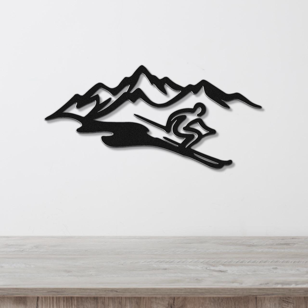 Black metal wall art featuring a skier silhouette in motion against a mountain range, mounted on a white wall above a wooden surface.