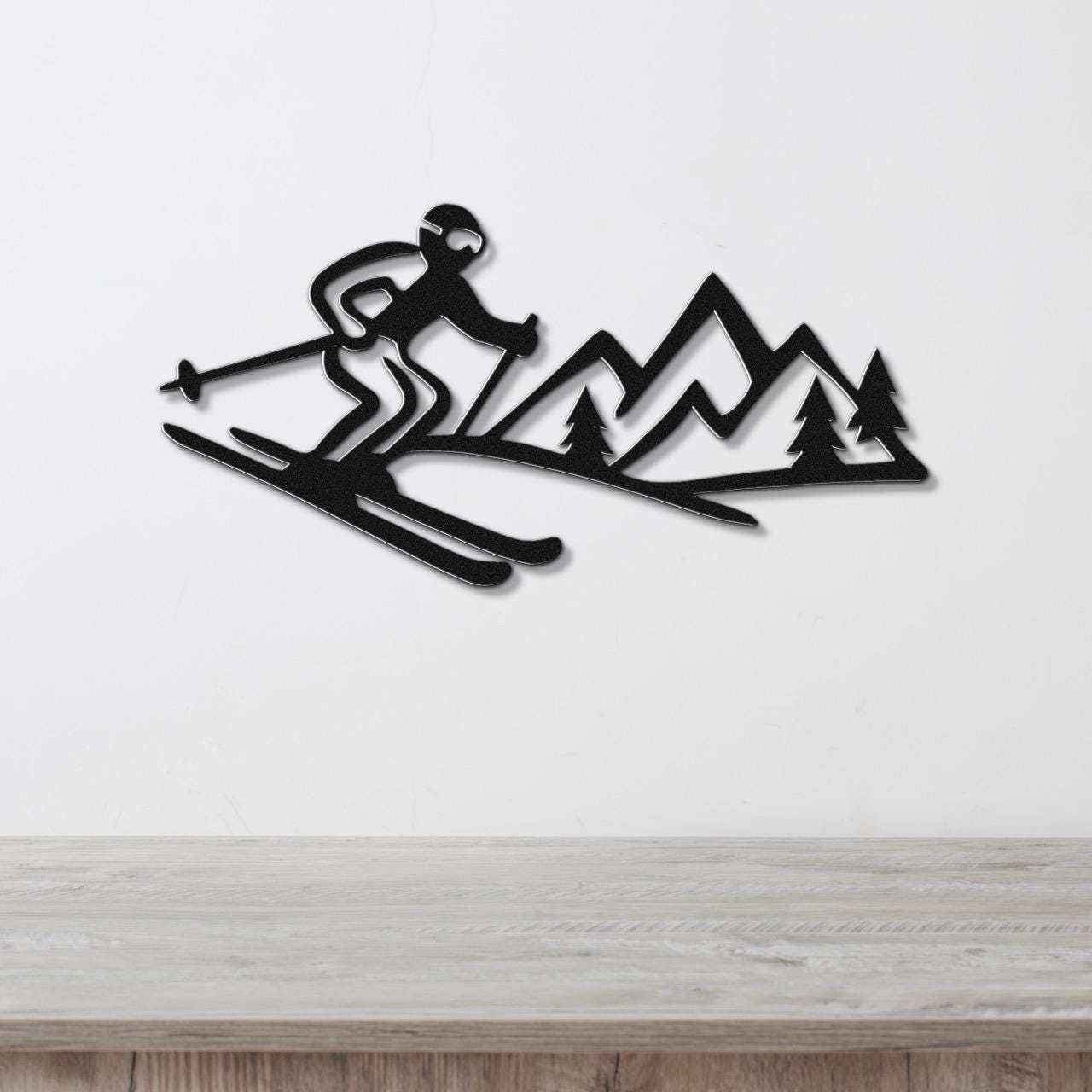 Black metal wall art cutout of a skier in motion with mountain and tree silhouettes, mounted on a white wall above a wooden surface.