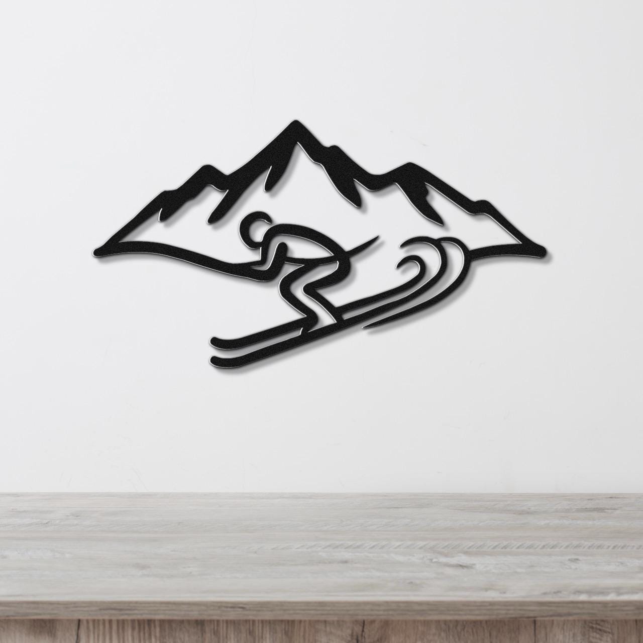 Black metal wall art featuring a skier silhouette in motion with mountain peaks, mounted on a white wall above a wooden surface.