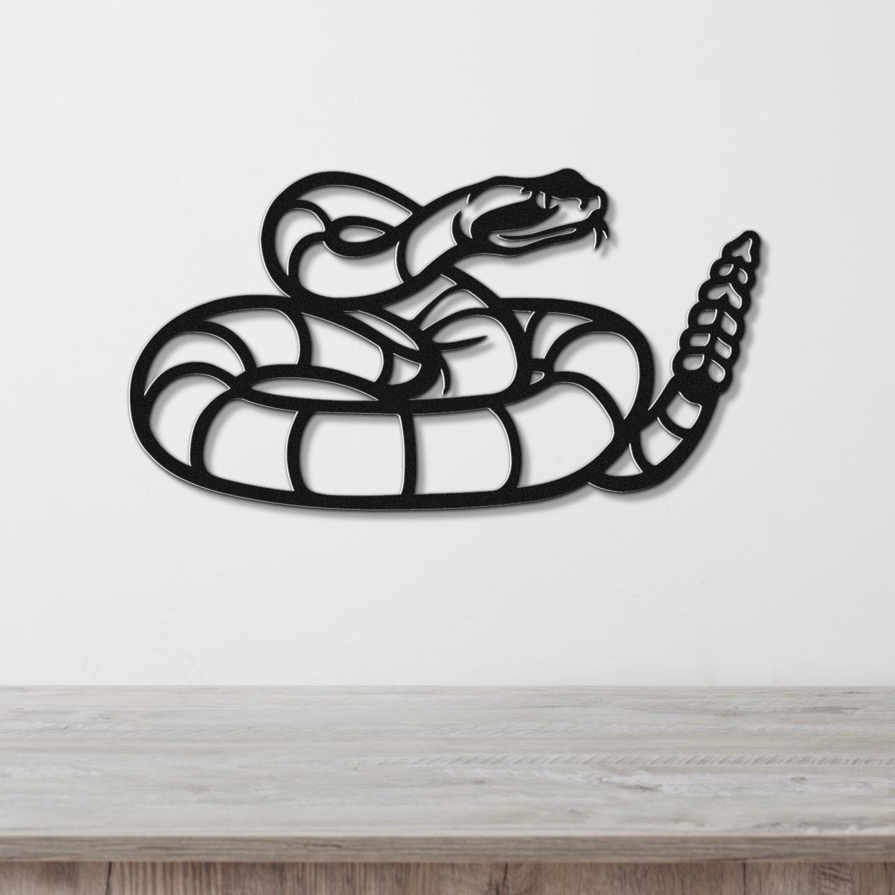 Black metal snake wall art with coiled body and raised head, featuring internal cutout details, mounted on a white wall above a wooden surface.