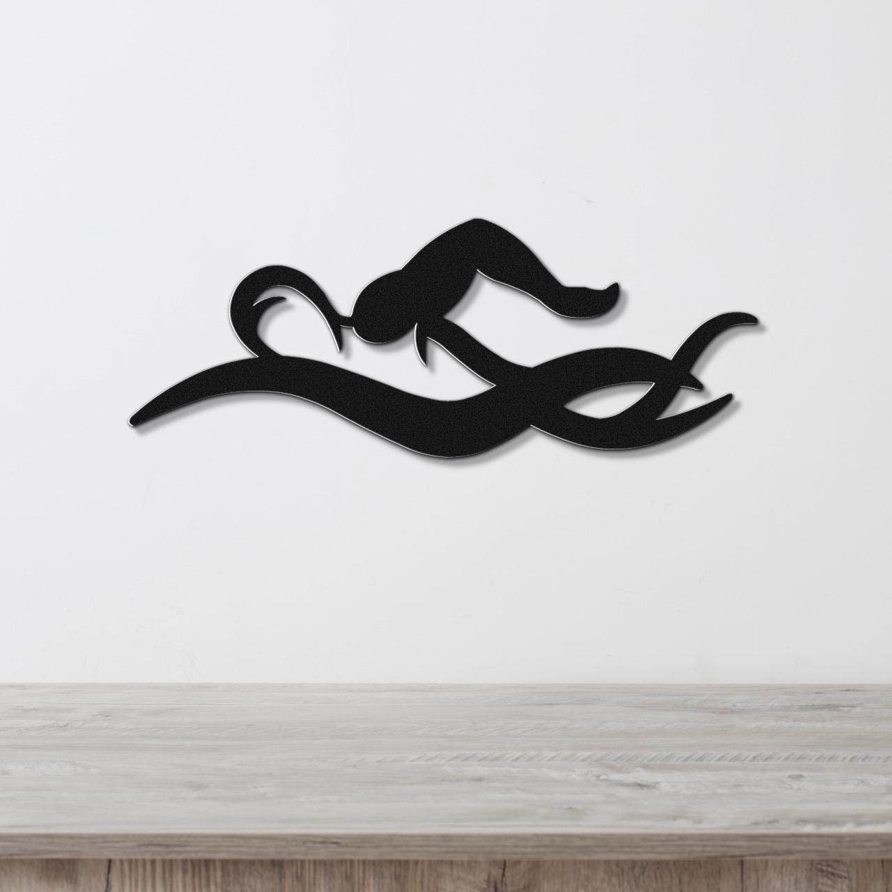 Black metal wall art of a swimmer in motion, with fluid cutout lines, mounted on a white wall above a wooden surface.