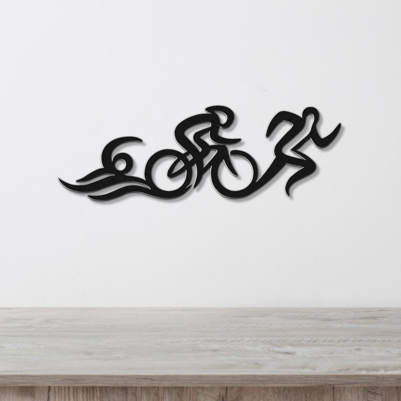 Black metal triathlon wall art with flowing cutout design of swim, bike, and run figures, mounted on a white wall above a wooden surface.