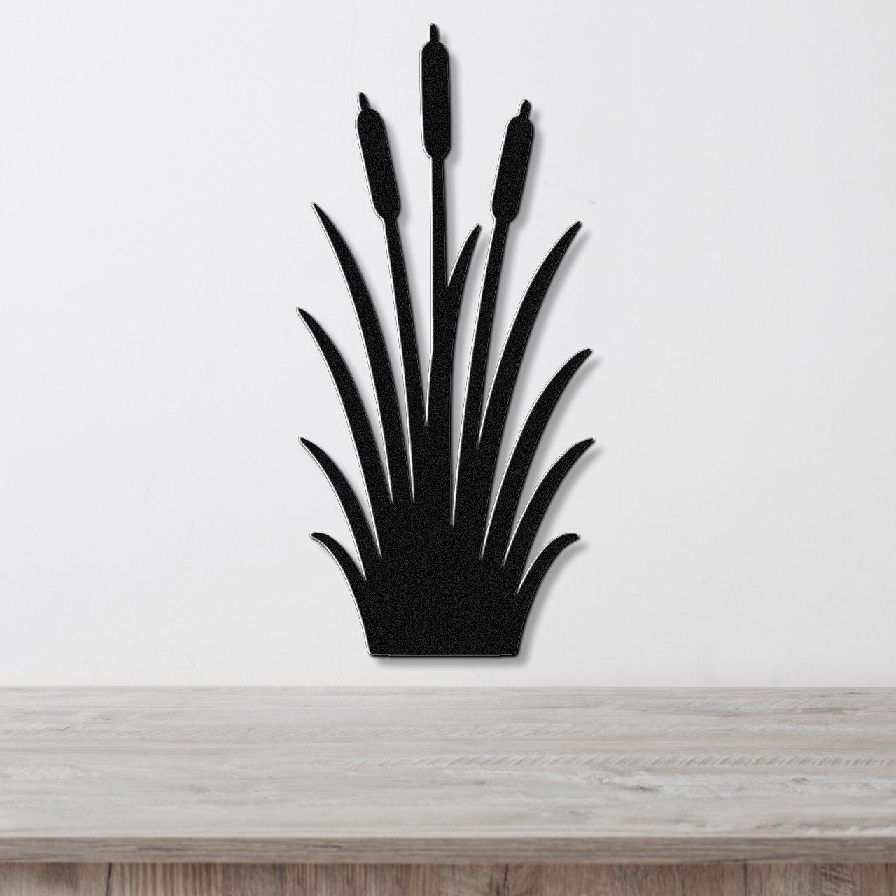 Black metal cattail wall art with a silhouette design of tall reeds and seed heads, mounted on a white wall above a light wood surface.