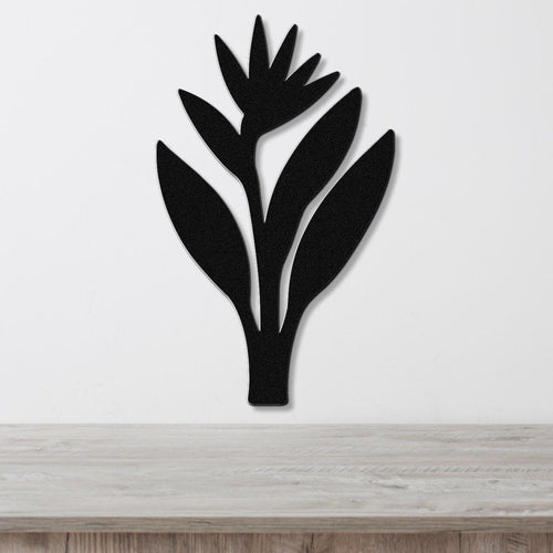 Black metal wall art in the silhouette of a Bird of Paradise plant with cutout leaves and flower, mounted on a white wall above a light wood surface.