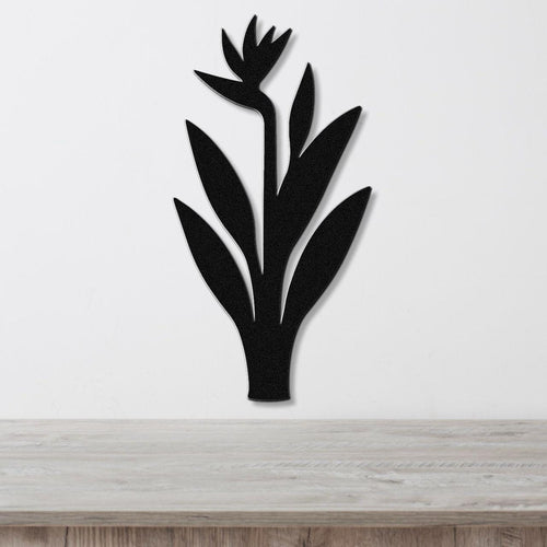 Black metal wall art in the silhouette of a bird of paradise plant with cutout leaves and flower, mounted on a white wall above a light wood surface.