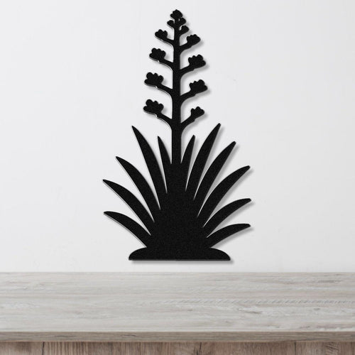 Black metal wall art in the silhouette of a blooming agave plant with a tall flower stalk and spiky leaves, mounted on a white wall above a wooden surface.