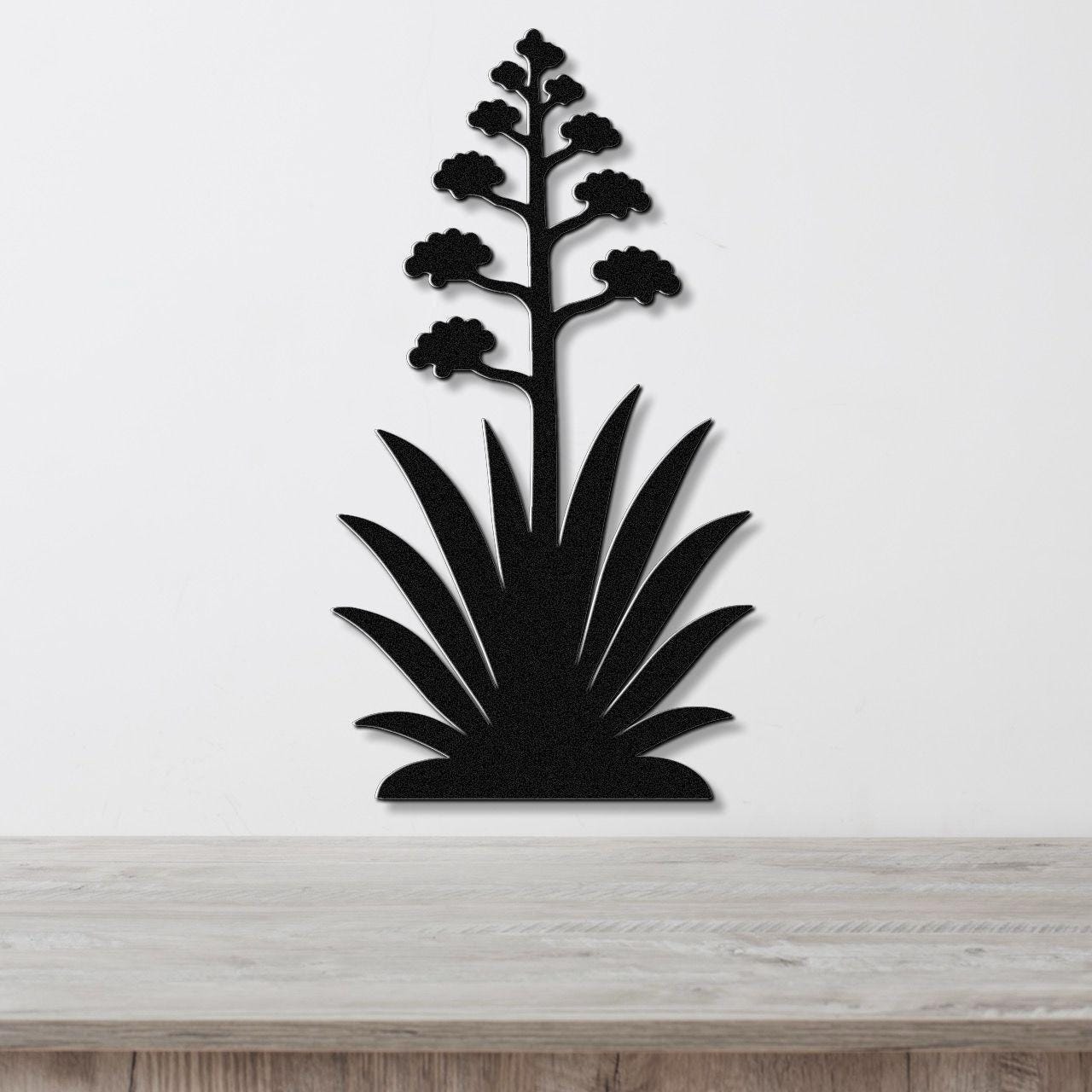 Black metal wall art in the silhouette of a blooming agave plant with a tall flower stalk and spiky leaves, mounted on a white wall above a wooden surface.
