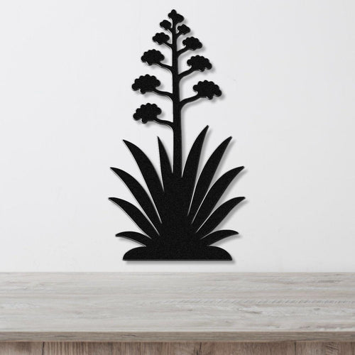 Black metal wall art in the silhouette of a blooming agave plant with a tall flower stalk and spiky leaves, mounted on a white wall above a wooden surface.