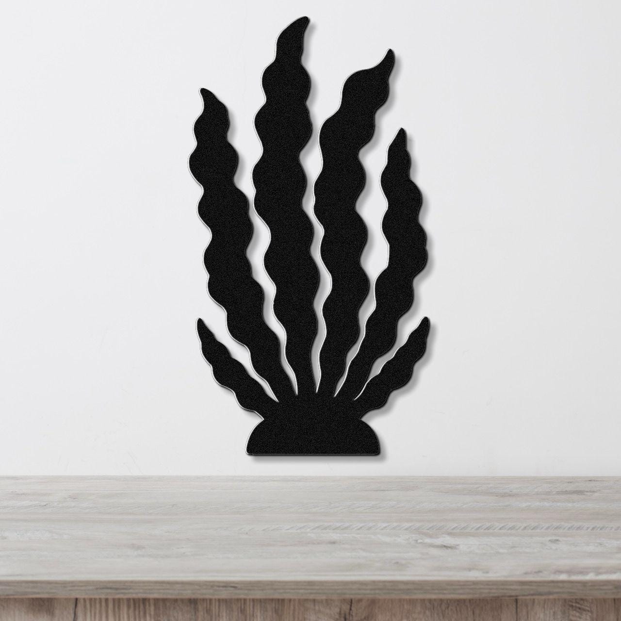 Black metal wall art in a kelp or seaweed silhouette design with wavy fronds, mounted on a white wall above a light wood surface.