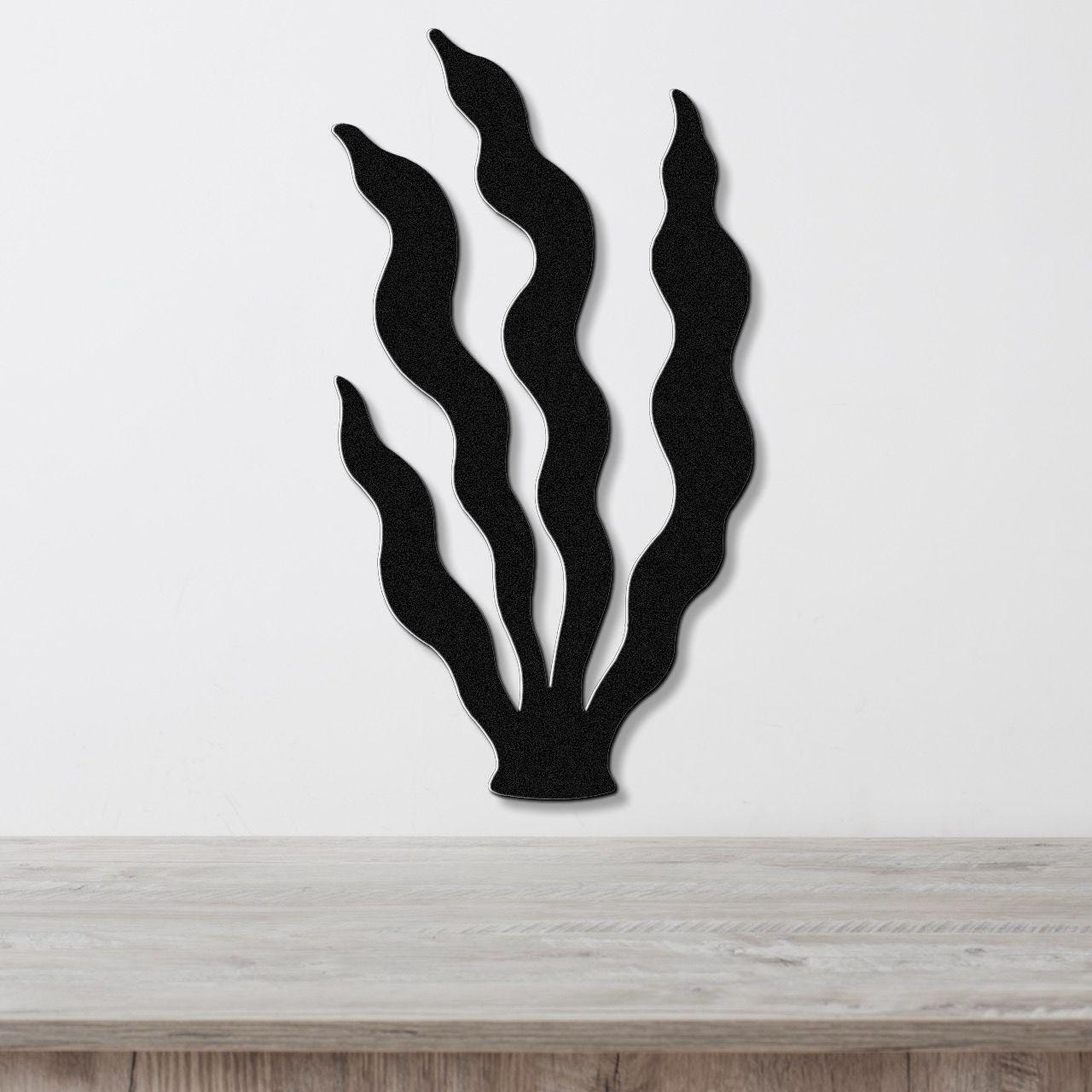 Black metal wall art in a kelp silhouette design with wavy fronds, mounted on a white wall above a light wood surface.