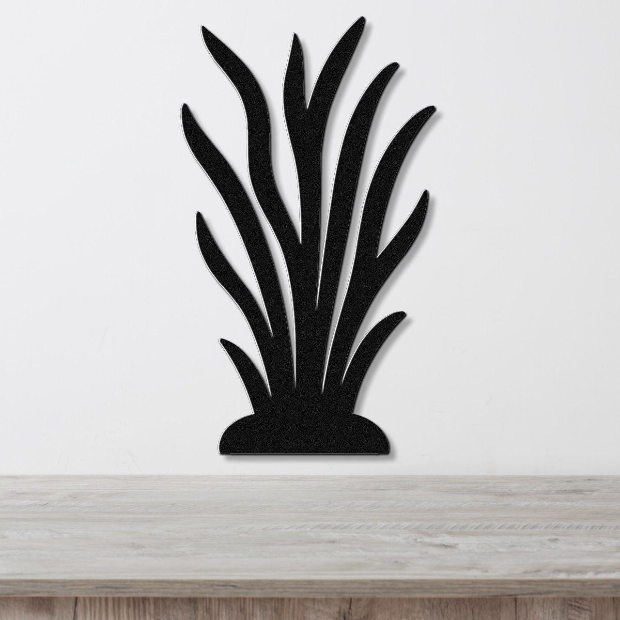 Black metal wall art in a kelp or seaweed silhouette design with layered leaf cutouts, mounted on a white wall above a light wood surface.