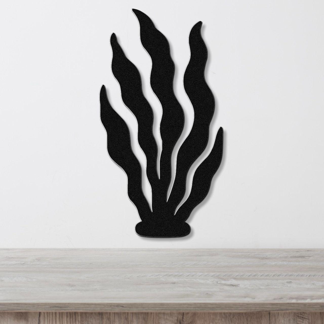 Black metal wall art in a kelp or seaweed silhouette with wavy leaf cutouts, mounted on a white wall above a light wood surface.