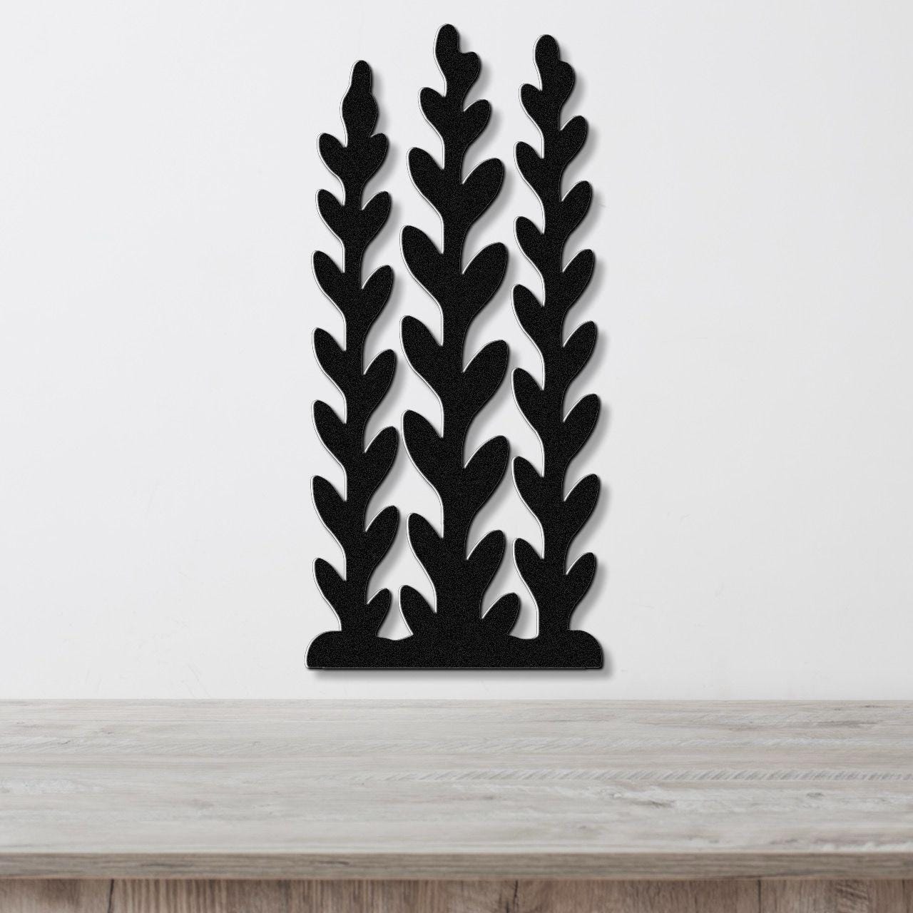 Black metal wall art with a kelp and seaweed silhouette design, featuring three vertical fronds with leaf-like cutouts, mounted on a white wall above a wooden surface.