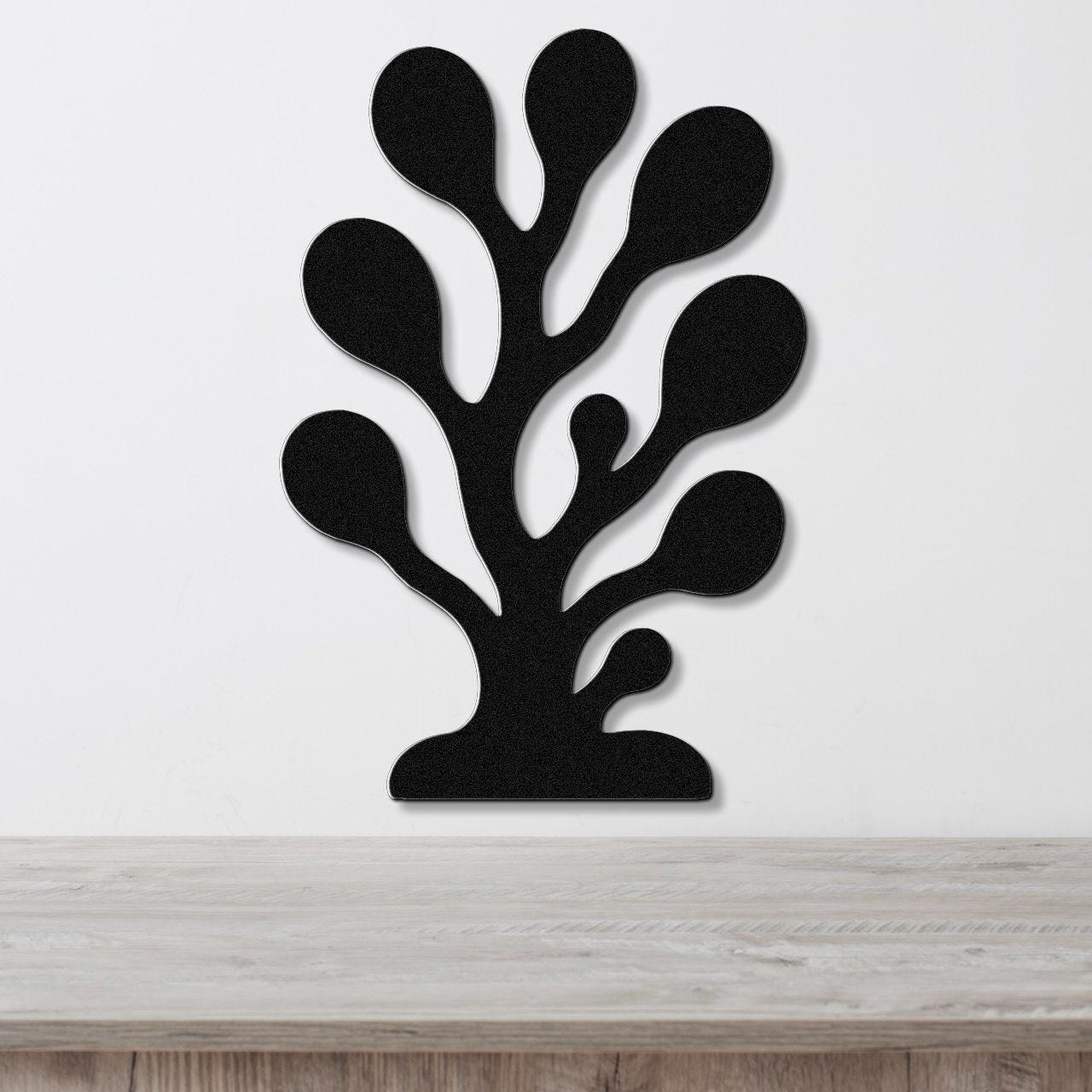 Black metal wall art in a coral or seaweed silhouette design with multiple rounded cutouts, mounted on a white wall above a light wood surface.