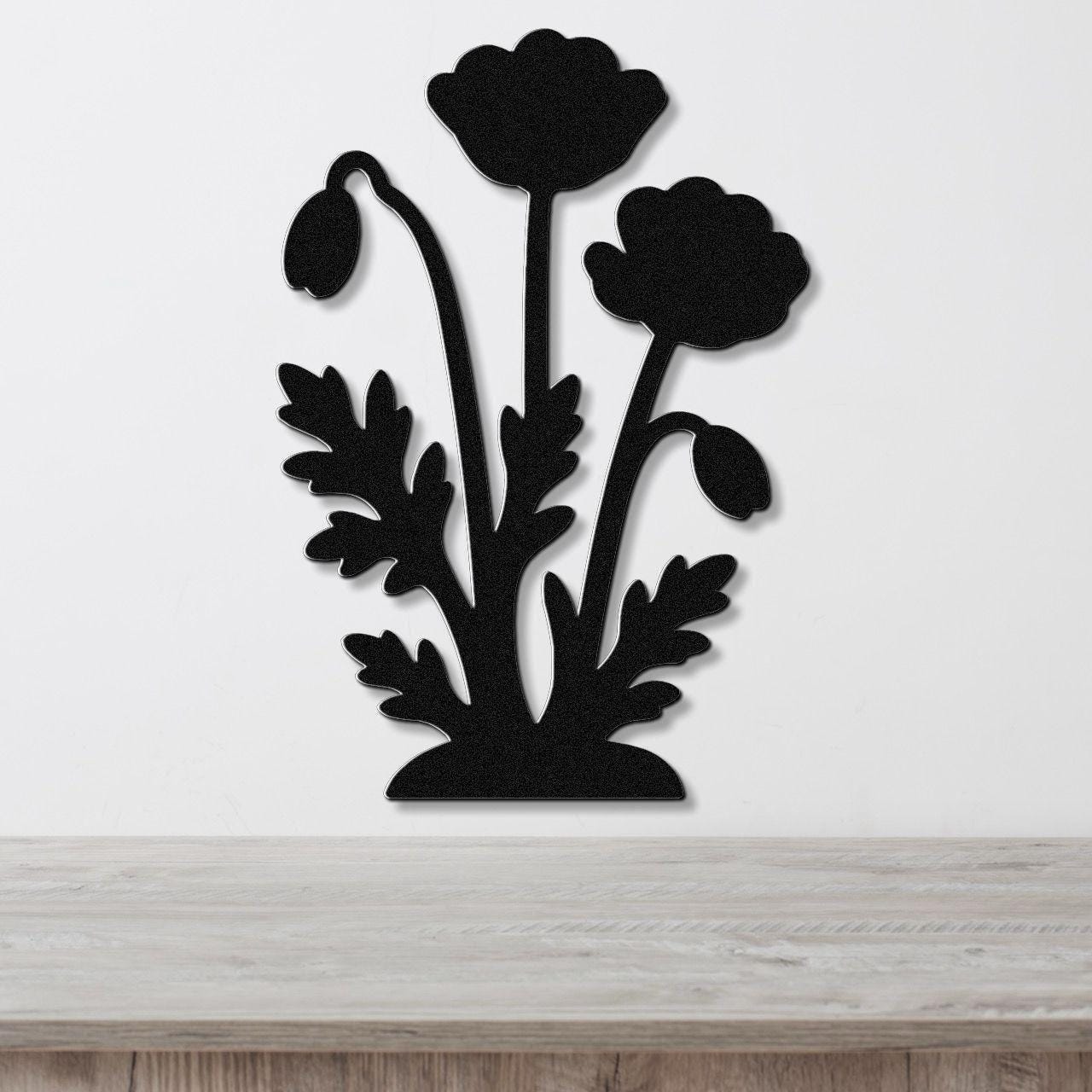 Black metal wall art featuring a silhouette of poppy flowers and leaves, mounted on a white wall above a wooden surface.