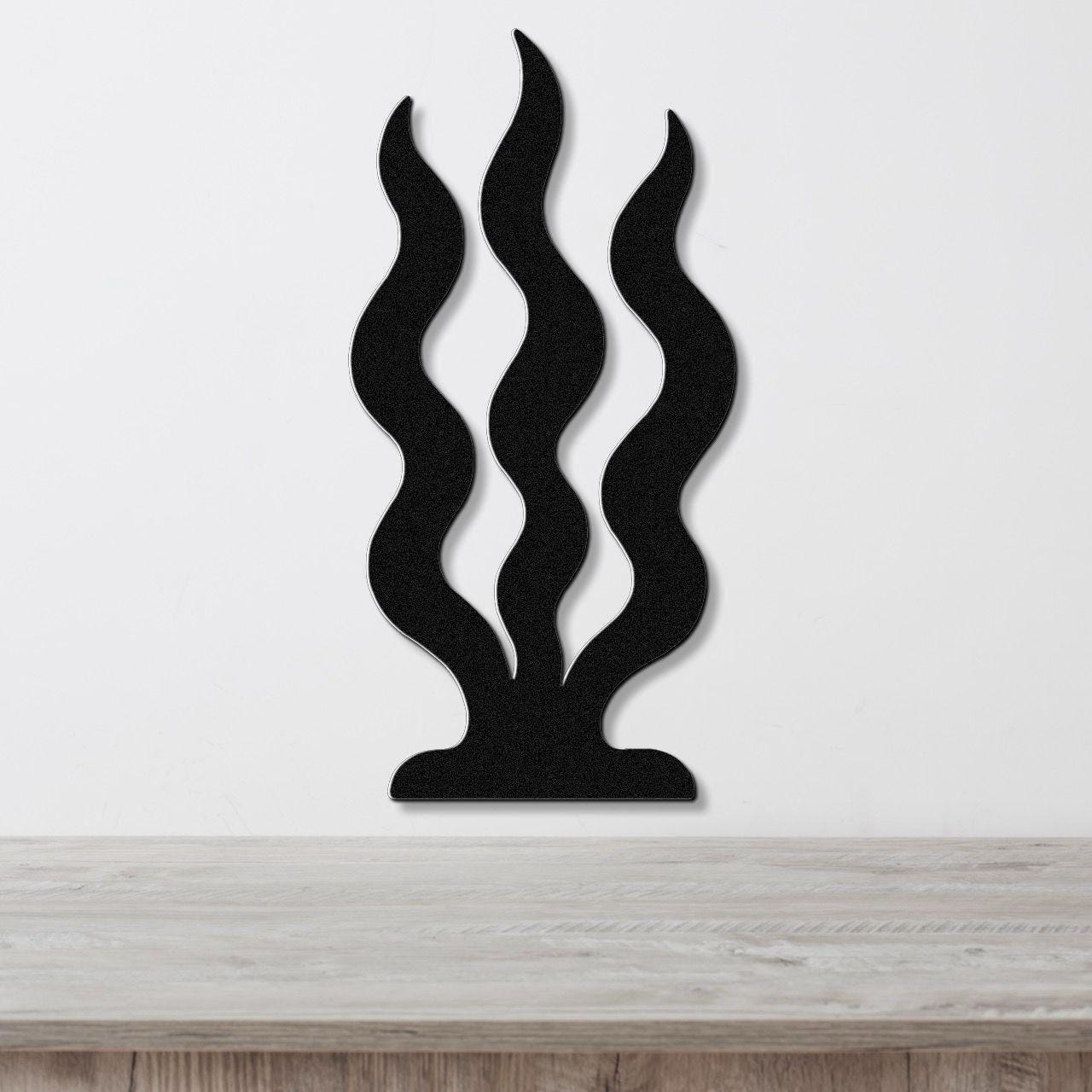 Black metal wall art with three wavy vertical cutouts resembling grass or seaweed, mounted on a white wall above a light wood surface.