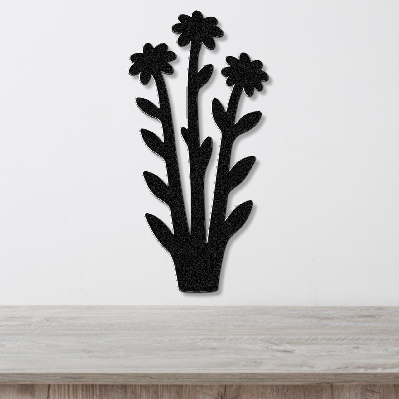 Black metal wall art featuring a silhouette of three wildflowers with leaves, mounted on a white wall above a wooden surface.
