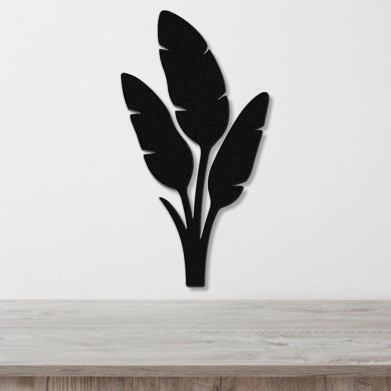 Black metal wall art in a tropical leaf silhouette design, mounted on a white wall above a light wood surface.