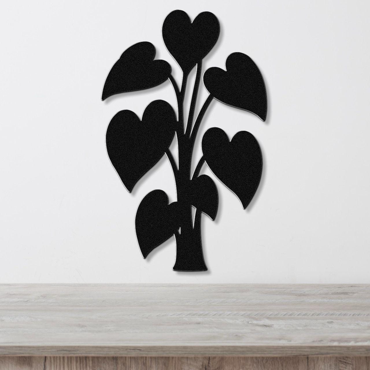Black metal wall art in the shape of a heart-leaf plant, mounted on a white wall above a light wood surface.
