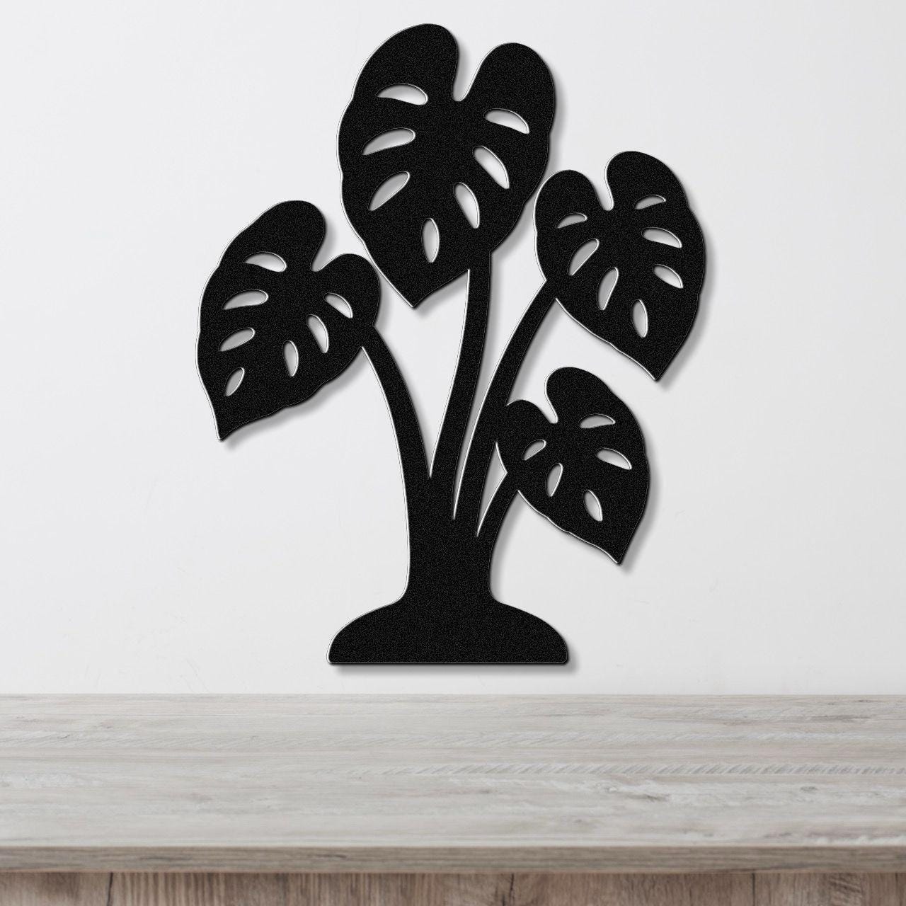 Black metal wall art cutout of a monstera plant with leaf perforations, mounted on a white wall above a wooden surface.