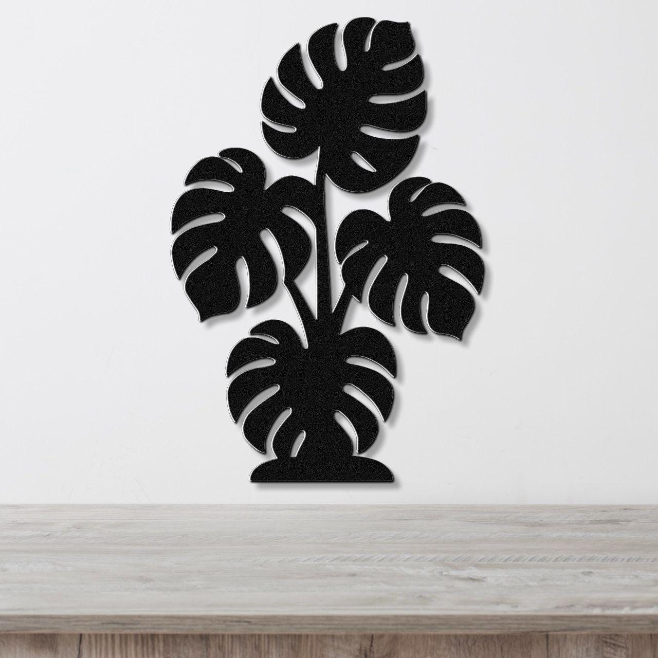 Black metal wall art cutout of a monstera plant with multiple leaves, mounted on a white wall above a wooden surface.