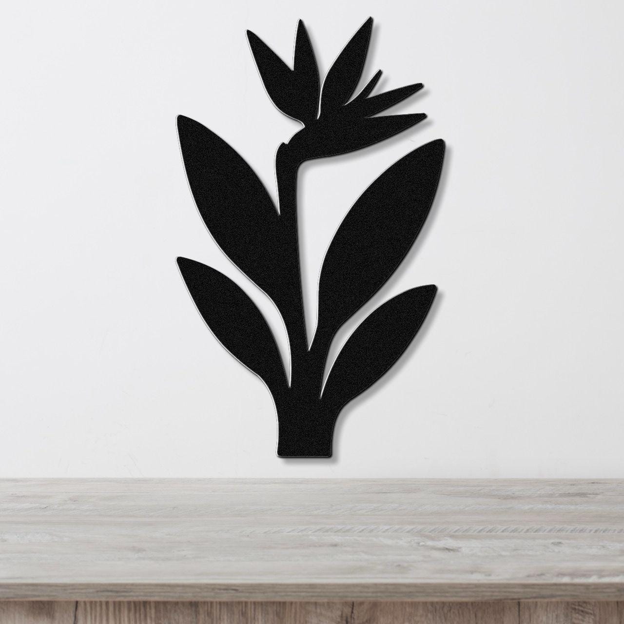 Black metal wall art in a clean Bird of Paradise silhouette with layered leaves and flower cutouts, mounted on a white wall above a light wood surface.
